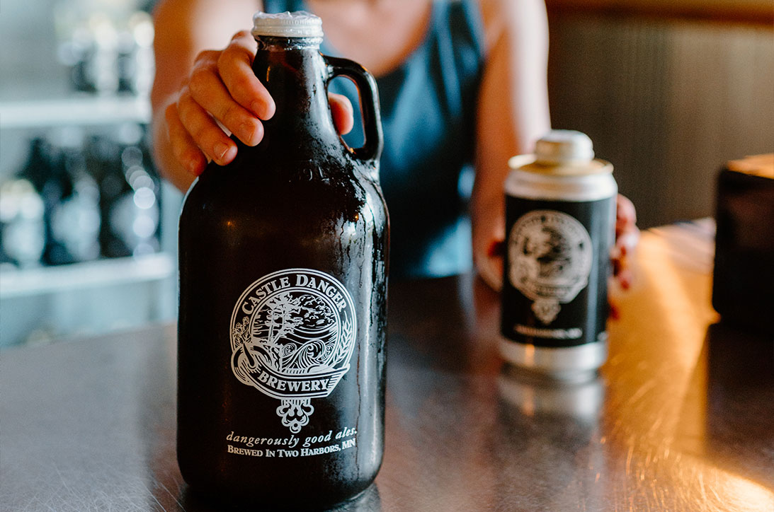 Person holding a growler and a crowler on a table.